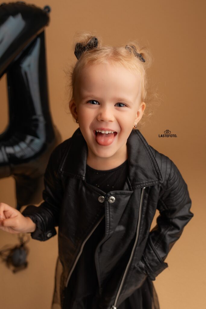 Kids Birthday Photoshoot in Studio – Why Capturing Every Age Matters 9 kids birthday photoshoot in studio – girl in a black leather jacket sticking out her tongue, black number 4 balloon on beige background