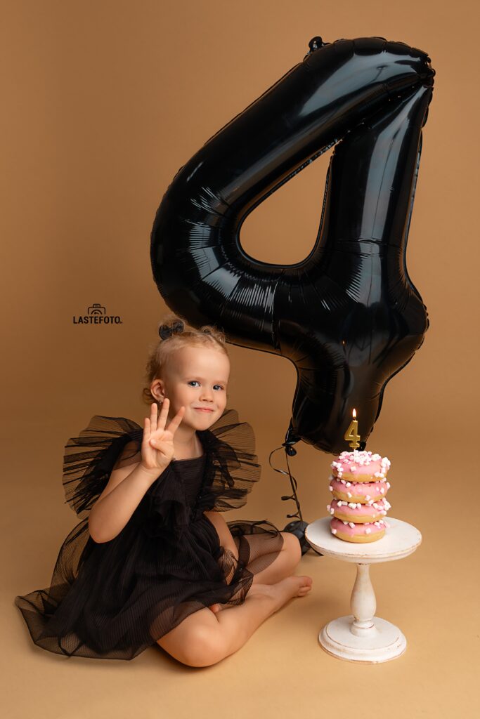 Kids Birthday Photoshoot in Studio – Why Capturing Every Age Matters 6 kids birthday photoshoot in studio – girl in black tulle dress sitting and showing four fingers; large black number 4 balloon and pink donut stack cake with candle