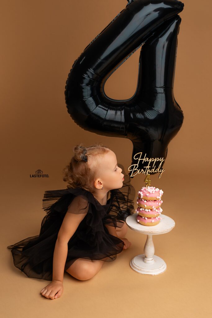 Kids Birthday Photoshoot in Studio – Why Capturing Every Age Matters 7 kids birthday photoshoot in studio – girl in black dress leaning toward a pink donut mini cake with “Happy Birthday” topper; large black number 4 balloon on beige