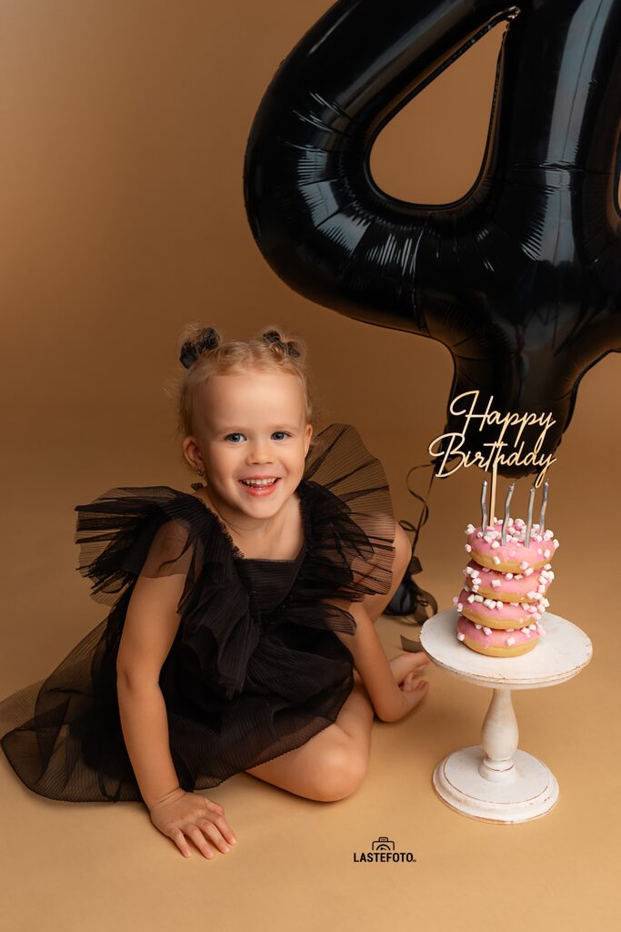 Kids Birthday Photoshoot in Studio – Why Capturing Every Age Matters 8 kids birthday photoshoot in studio – smiling girl in a black tulle dress with a large black number 4 balloon and a pink donut mini cake with candles on a beige background