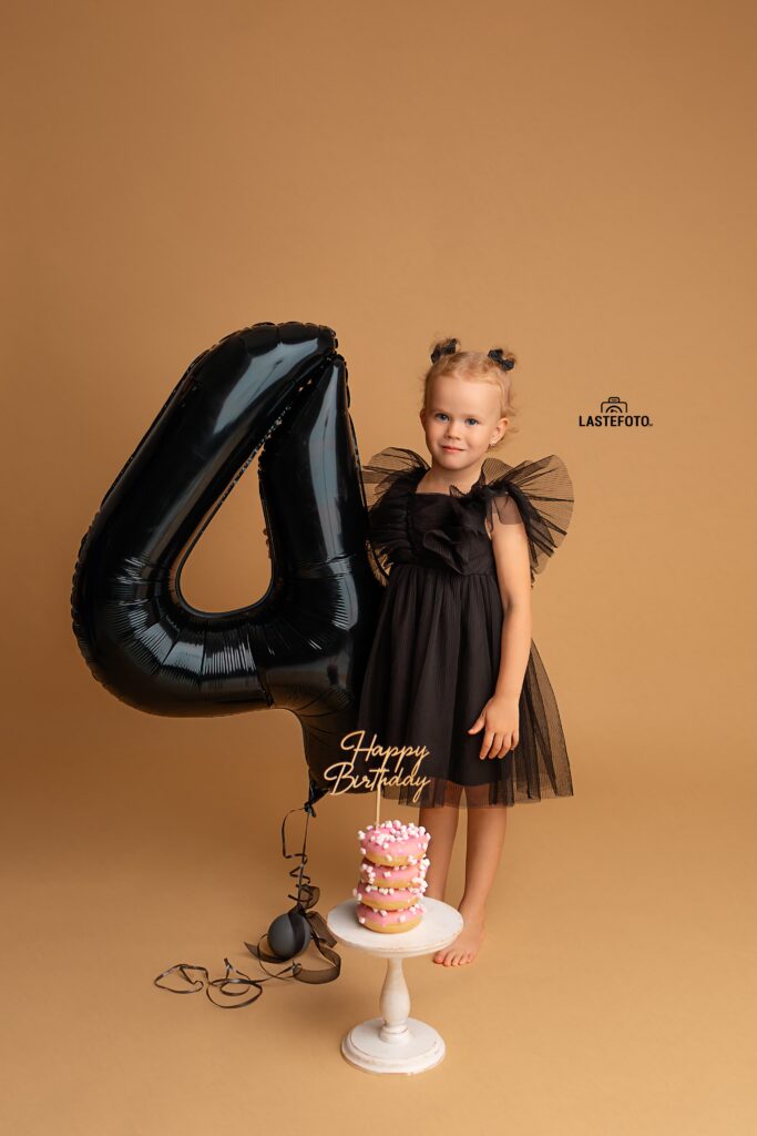 Kids Birthday Photoshoot in Studio – Why Capturing Every Age Matters 2 kids birthday photoshoot in studio – girl in a black dress standing beside a large black number 4 balloon; pink donut mini cake with “Happy Birthday” topper on a stand, beige background