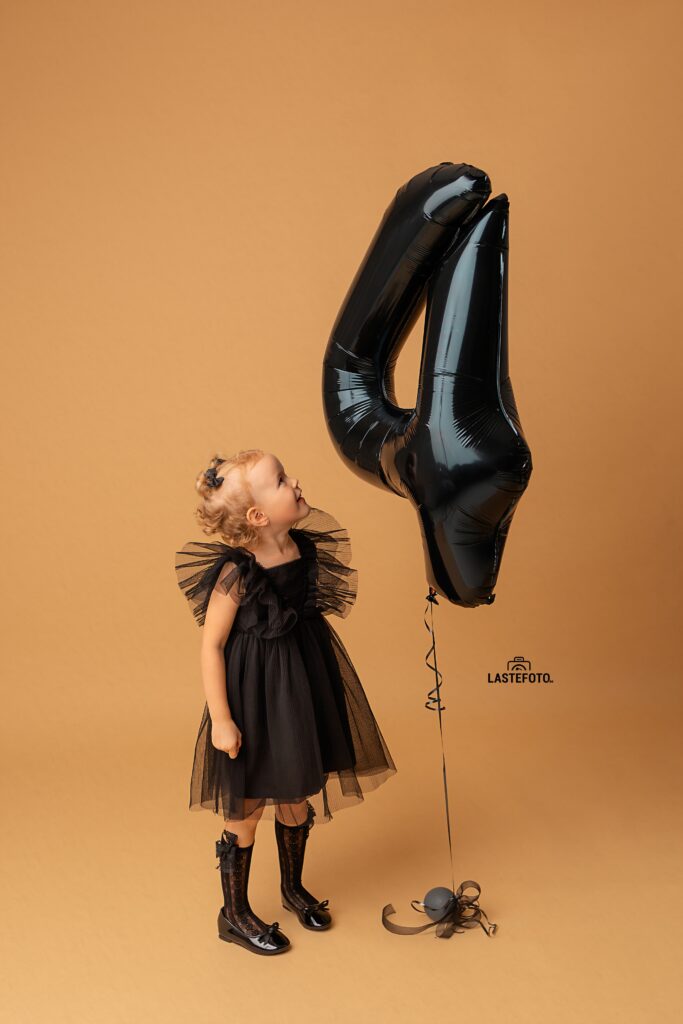 Kids Birthday Photoshoot in Studio – Why Capturing Every Age Matters 4 kids birthday photoshoot in studio – girl in a black dress looking up at a large black number 4 balloon on a beige background