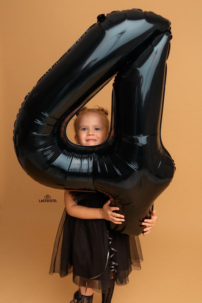 Kids Birthday Photoshoot in Studio – Why Capturing Every Age Matters 3 kids birthday photoshoot in studio – girl peeking through a large black number 4 balloon, black dress on a beige background