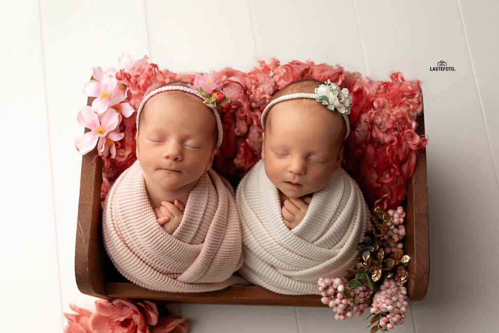 Newborn Twins Photoshoot: 6 Practical Tips for Calm, Confident Planning and Preparation 7 Newborn Twins Photoshoot: two sleeping twin girls swaddled in blush and cream, lying side by side in a wooden baby bed with floral headbands and a pink wool backdrop