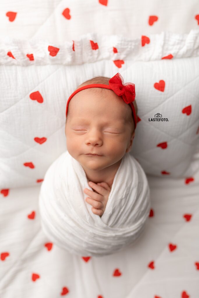 Newborn Twins Photoshoot: 6 Practical Tips for Calm, Confident Planning and Preparation 8 Newborn Twins Photoshoot: sleeping baby girl in a white wrap with a red bow headband, close-up on a white blanket with red hearts