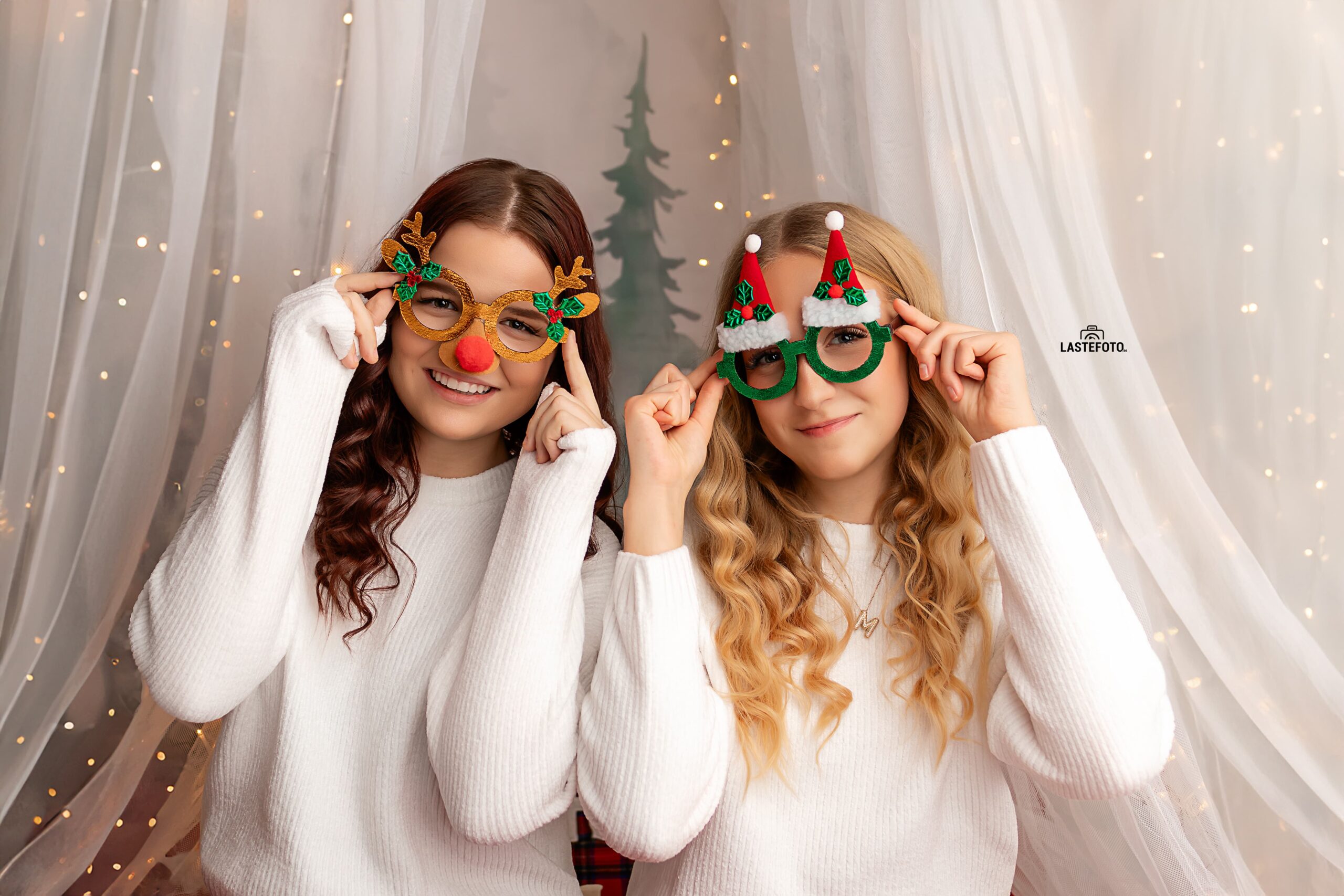 Christmas Photo Session in Tallinn – two teenage girls in white sweaters and festive glasses smiling in a cozy studio with fairy lights