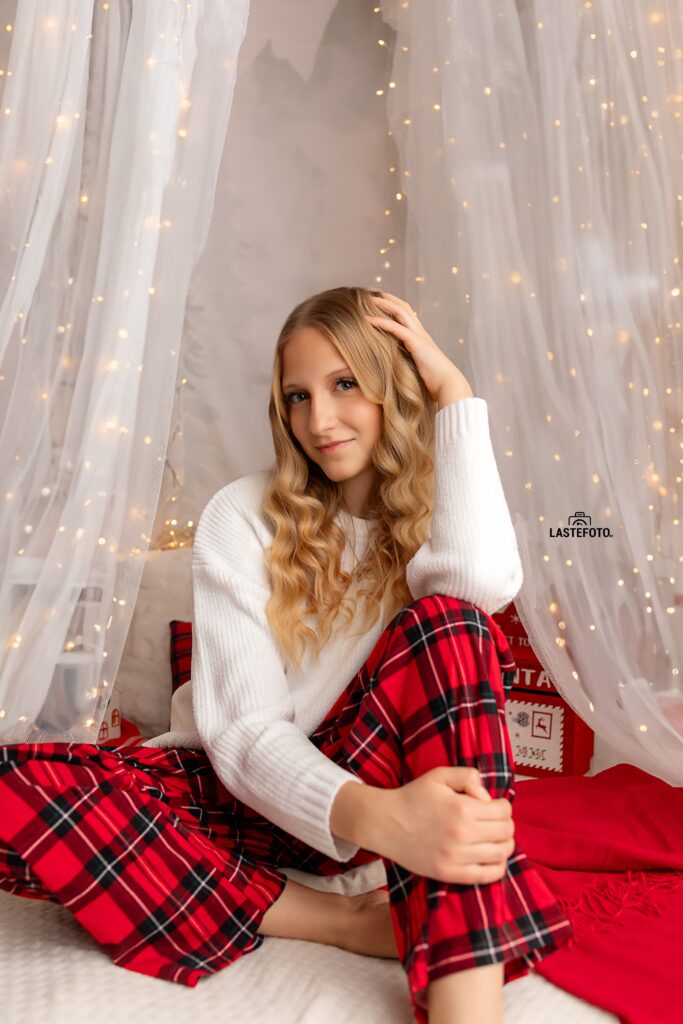 Christmas Photo Session in Tallinn – girl in white sweater and red plaid pants sitting on a bed in a bright studio with fairy lights