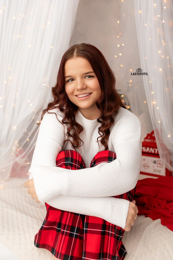 Christmas Photo Session in Tallinn – girl in white sweater and red plaid skirt sitting in a bright studio with fairy lights and festive decor