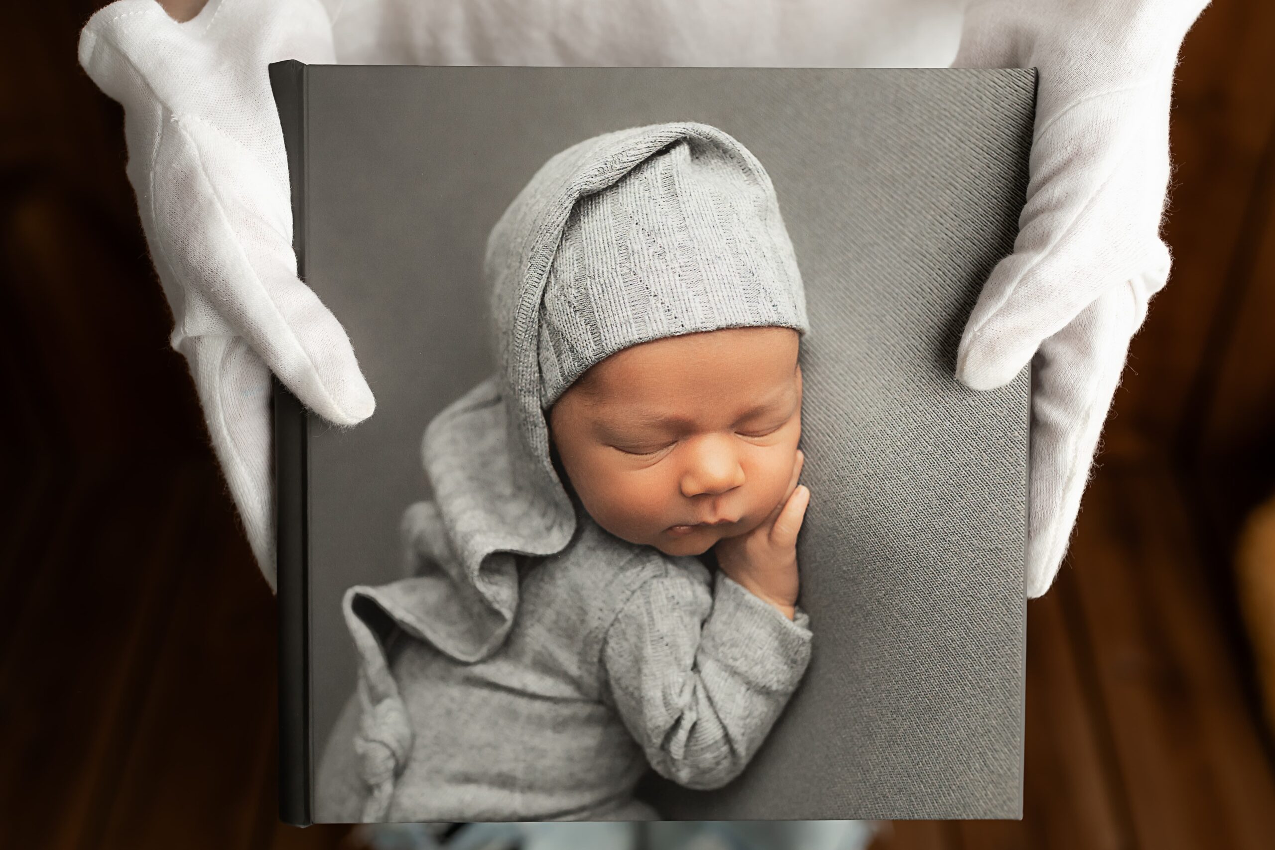 Photobook With Your Heartwarming Family Story - Made to Be Opened Again and Again 1 Photobook with a sleeping newborn in a gray hat on the cover, held carefully in white gloves.