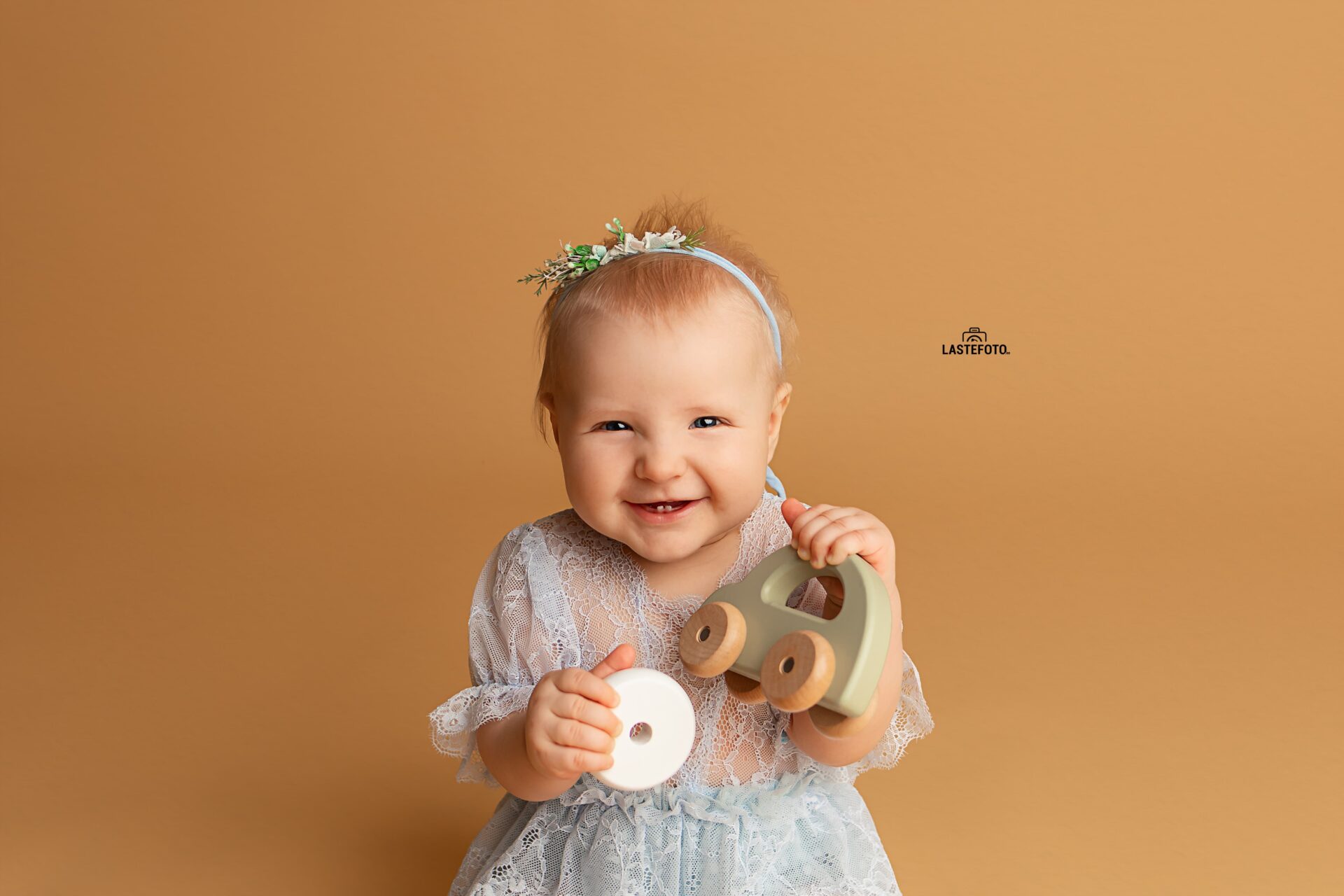 best age for baby photos – smiling baby girl in a light blue lace dress holding a wooden toy car against a warm brown background