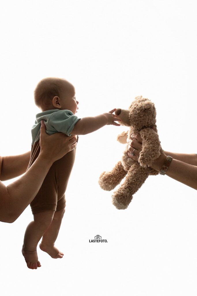 Best Age for Baby Photos: What to Expect at 4, 6, 9 and 11 Months 14 best age for baby photos – baby reaching towards a teddy bear held by adults against a bright background