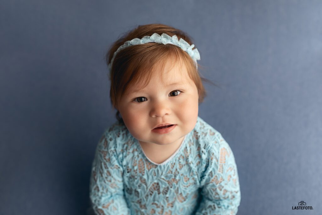 Best Age for Baby Photos: What to Expect at 4, 6, 9 and 11 Months 8 best age for baby photos – baby girl in a light blue lace outfit and headband sitting against a blue background and looking at the camera