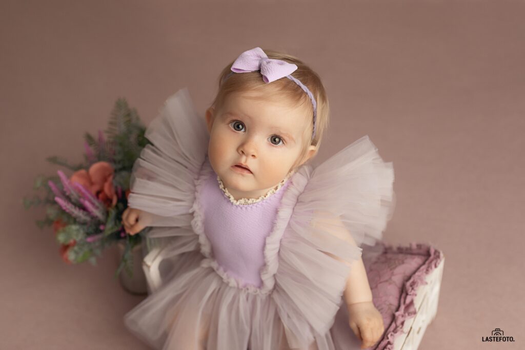 Best Age for Baby Photos: What to Expect at 4, 6, 9 and 11 Months 9 best age for baby photos – baby girl in a lilac dress with full tulle sleeves sitting beside flowers against a pink background